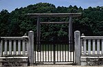 Torii gate behind a concrete fence. In the back behind the torii there is water and behind it trees.