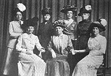 Seven white women (three seated, four standing) posed for a group photograph. They are all wearing long skirts and large hats. Several are wearing suit jackets.