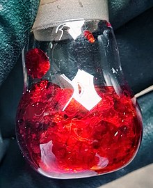 A bright red substance in a small glass flask, held by gloved fingers