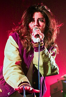 A young brunette woman wearing a baseball-style jacket, singing into a microphone against a pinkish background.