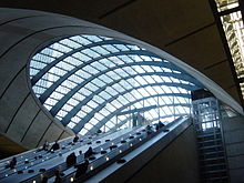 Escalators descending into an underground station with a glazed canopy above, with a glazed lift shaft on the side