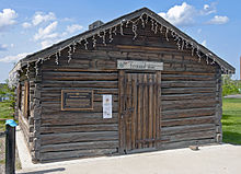 A dark brown log cabin, topped with a pointed roof, seen from the front. Over its main door is a sign saying "Fireweed Studios" in script on white with floral decoration. To its left is a bronze metallic plaque with text explaining the building's history.