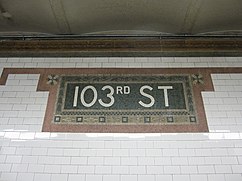 A mosaic name tablet on the wall of the 103rd Street station. There are white ceramic tiles surrounding a tablet with bronze, salmon, and green tiles. At the center are white mosaic tiles spelling out "103rd Street".