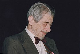 A quarter-profile photograph of Gunnar Dybwad. He has grey hair laid over to the photographer's side and a grey Van Dyke beard. He has a deeply lined face and is looking slightly down. He is wearing a suite and a maroon bow tie. The background is totally black.