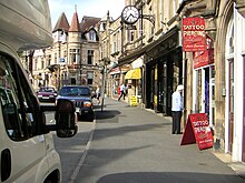 Single carriageway road on a slight curve stretching into the distance lined with stone-fronted buildings used by shops and a hotel on the right, with a parked motorhome and car to the left