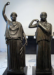 Two black bronze statues of women; one has her right hand raised above her head, the other adjusts her clothing.