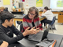 A student gestures with his hands out in front of a computer to a female teacher.