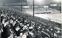 A black and white photograph of a ballpark's grandstand filled with people during a baseball game.