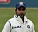 A man in the white Indian test cricket jersey, wearing a blue hat. In the background is the field of a cricket ground.