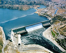 Arial photograph of an L-shaped concrete hydro-electric dam set against road swerving in a green landscape. The center of the damn is colored white as water rushes down the spillway.