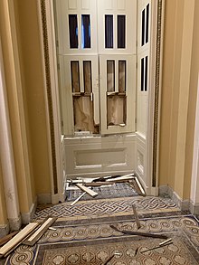 A photo taken from the inside of the Capitol building. Windows are broken, along with their wooden frames. They are boarded up.