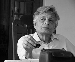 black-and-white image of Irfan Habib wearing a white shirt, sitting at a desk with a binder clip in right hand, looking right of camera