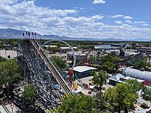 The lift hill from an opposing angle is seen, with several flat rides in the background. The viewpoint is in the air, with the ascent of the lift hill on the left and the out and back portion of track visible. Several buildings can be seen on the right side, with the Fire Ball and Downdraft flat rides hugging the background of the lift hill. Trees and housing can be observed in the distance with mountains.