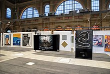 A distant view of Burak Karavit's work on display from the Zurich Train Station. Plastic Dreams hangs in the middle of the train station. There are also works by other artists next to and behind his work.