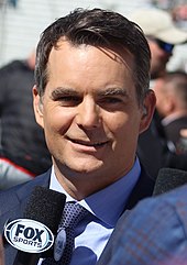 A smiling Jeff Gordon, in a suit in front of a Fox Sports microphone