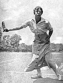 A white woman playing tennis, wearing a long buttoned skirt and a short-sleeved blouse.