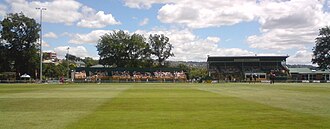 A cricket ground with two stands and several trees visible.