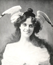 A young smiling white woman with voluminous dark hair, wearing a hat with two full bird wings attached at the sides