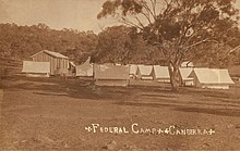 Single wooden cabin and many white tents in open dusty field with single tree in foreground