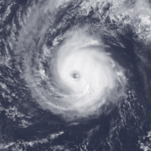A satellite image of a hurricane over the Central Pacific Ocean. It has a clear but ragged eye surrounded by deep but somewhat lopsided convection which is mostly concentrated in the northeastern quadrant; an arc of thin high clouds begins on the system's south side, curving around the western flank.