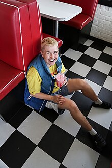 Alex the Astronaut sitting on the floor of a diner, drinking a milkshake. They have short blond hair and a big smile on their face.