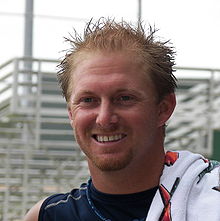 A smiling young man with short, spiked red hair and a towel laid over his left shoulder