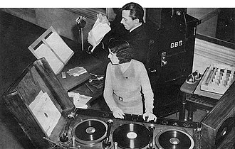 Black-and-white photograph, view from above, of a woman facing forward, but with her face turned to our left, and a man facing left behind her. They are in a room with sound equipment carrying a CBS label, with three turntables in front of the woman, and some scripts on lecterns at the left.