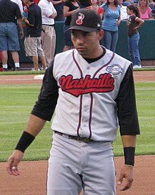 A man with a black cap, gray baseball jersey with black sleeves and "Nashivlle" written across the chest in red, and gray pants stands on a baseball field.