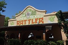 The entrance of the New Mexico Rattler's entrance is observed from a ground level. The logo of the roller coaster is depicted on a tan façade, with the addition of green cacti on either side. Foliage surrounds the lower half and background of the ride's entrance.
