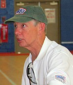 An older Caucasian man wearing a dirty green baseball cap and white polo shirt is sitting down on a chair in the middle of a basketball court. He is looking off to the left while conversing with a person off-camera.