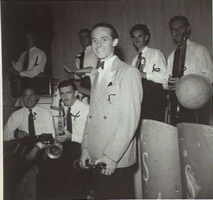 Smiling man standing in front of six other men – all holding musical instruments.
