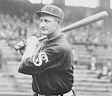 A man in a dark baseball uniform and cap poses with a baseball bat on his shoulder.