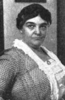 A middle-aged white woman with dark hair braided across the crown, wearing a print dress with a white ruffled front section and collar