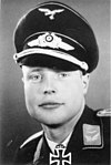 A man wearing a military uniform, peaked cap, and an Iron Cross displayed at the front of his uniform collar.