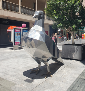 Photo of a two-meter tall, polished stainless steel sculpture of a pigeon installed in an outside area of a shopping mall