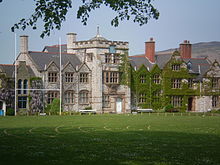 A stone building, partly ivy-covered, seen from a slight angle, with a playing field in the foreground. The front of the building contains a square battlemented tower, two shaped gables and a series of plain gables.