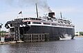 A large black ship with white upper decks rests at a berth, its rear surrounded by wooden posts. The rear access to its vehicle decks is opened upward.