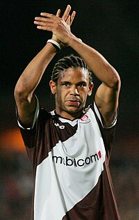 African man in a diagonally striped brown-and-white jersey with a sponsor logo across the chest. His hair is cornrowed, and his arms are crossed above his head.