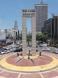 A marble monument, monolithic in structure, situated on a traffic island painted red and yellow. The monument has text near its top which says: "WELCOME QUEZON CITY"