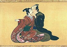 A shunga-style scroll on skill. The background is yellow, and a partially shaved, middle aged men is sitting in green and black robes. Next to him and halfway on his knees is an adolescent boy in an ornate red dress. The two are kissing on the lips with eyes closed; the older man is holding the youth's chin, and the boy's bare feet are exposed.