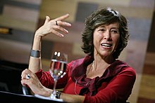 Maria Rita Kehl sitting at a table, in an event, talking ang gesturing with her hands. On the table is a glass of water
