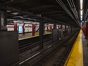 View from the southbound platform of the 47th–50th Streets–Rockefeller Center station. There is a yellow tactile strip on the platform's edge. The southbound local and northbound express tracks are separated by a set of columns. In the background is the northbound local track, adjacent to a white tiled wall with a red-tile band.