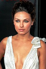 A woman with her hair tied back, poses while wearing a white dress that slightly exposes her breasts.