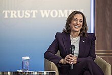 A woman in a purple suit jacket smiles, holding a wireless microphone with both hands while sitting relaxed in a chair, with the text "TRUST WOMEN" on a screen behind her.