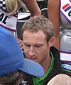 A man in his late twenties is looking down from the camera and signing autographs for others. He is wearing a black fleece with sponsors logos and a green T-shirt