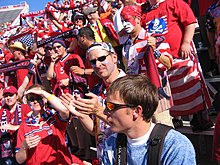 A number of American fans, dressed in red, stand in bleachers holding United States paraphernalia.