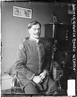Black and white photographic portrait of Sarath Grosh, indian prince, sitting in a chair in a room in Chicago. Picture has text written in write color at the right edge, saying: "Prince Sarath Ghosh of India"