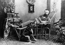Franz Benque posing in a wooden chair with his legs up, 1883