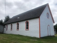 Photo of a single storey wooden building, painted white with orange trim. It has a circular window and an arched door in the gable end, and arched windows along the side facing the viewer.