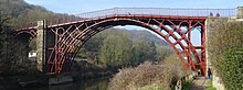 Red painted skeleton high arched metal structure with masonry pillars at each side spanning a deep gorge with wooded sides